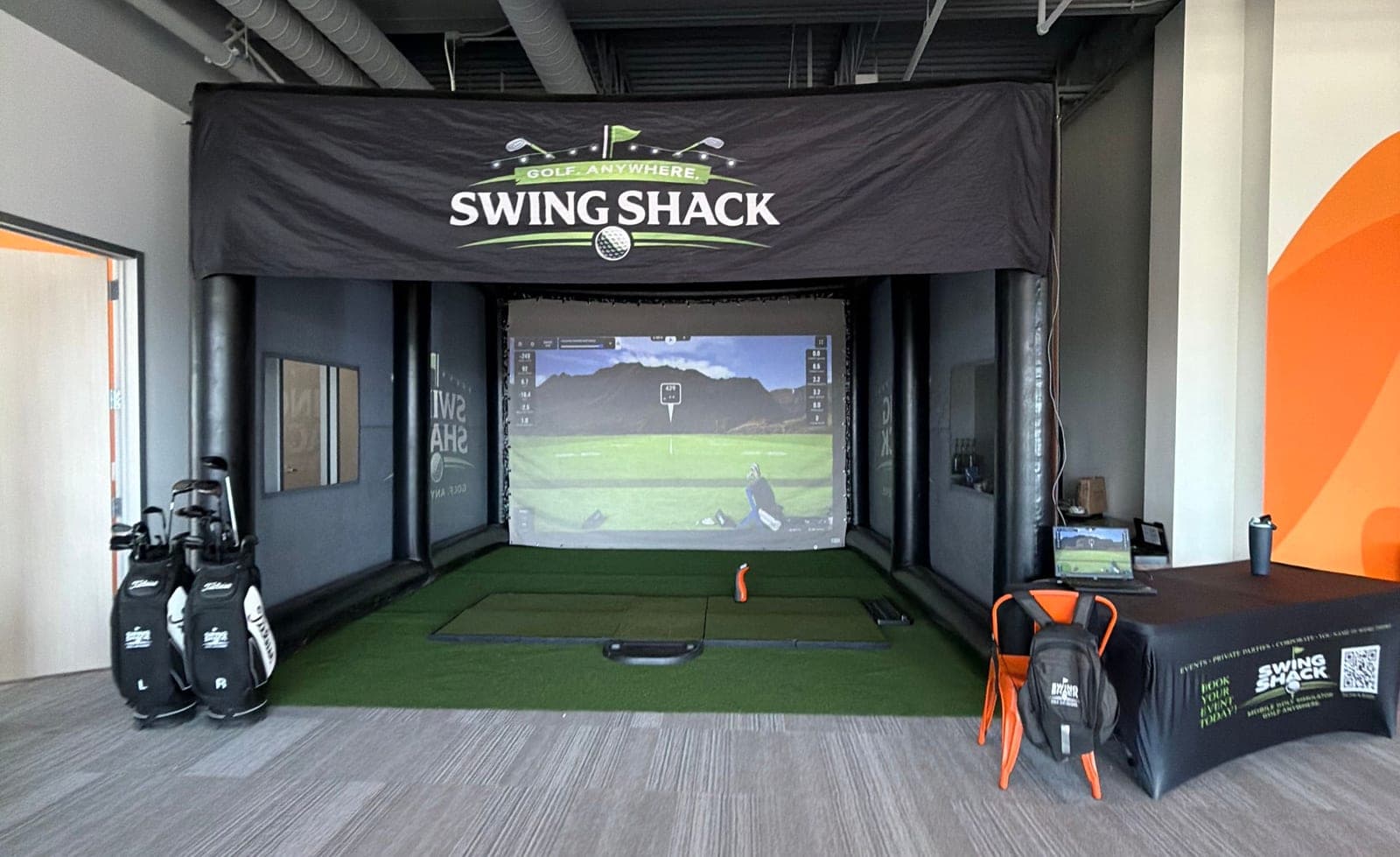 Swing Shack simulator set up indoors at an office with a bright course on screen, golf bags, and a branded table
