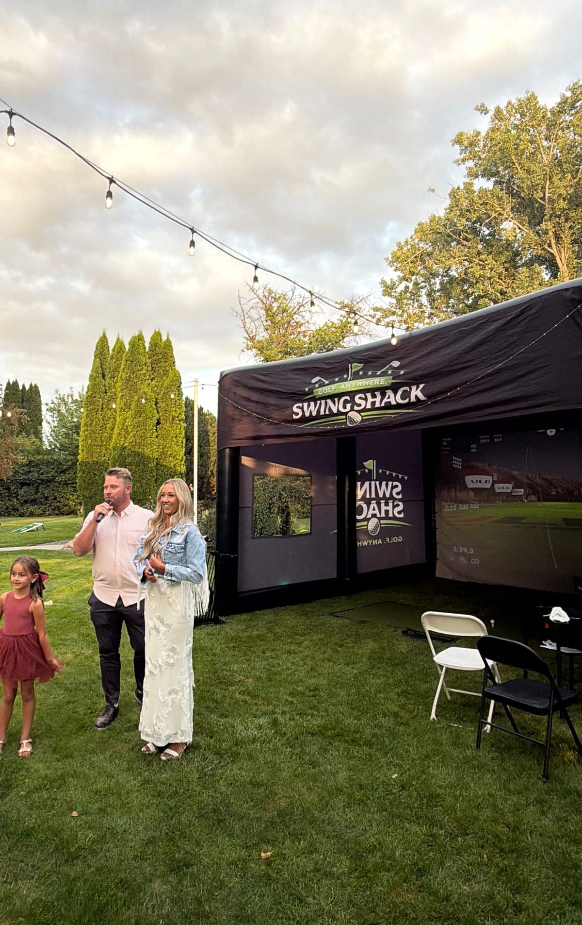 A newlywed couple giving a toast on the lawn beside the Swing Shack simulator at a backyard wedding